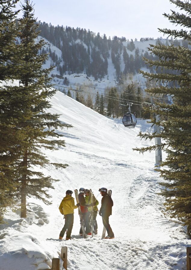 Four people with skis stand together on a snowy slope near trees, with a ski lift and gondola visible in the background on a sunny day—capturing the spirit of Aspen travel and Rocky Mountain hotels.