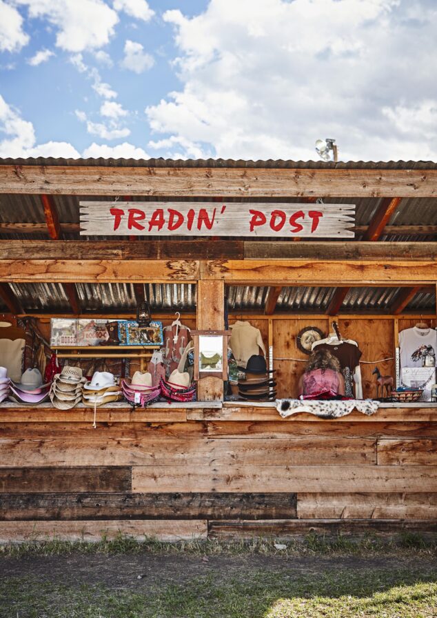 An editorial capture of wooden trading post booth displays cowboy hats, shirts, and various goods under a corrugated metal roof with a sign reading "TRADIN' POST," reminiscent of frontier stops in the Rocky Mountains of North America.