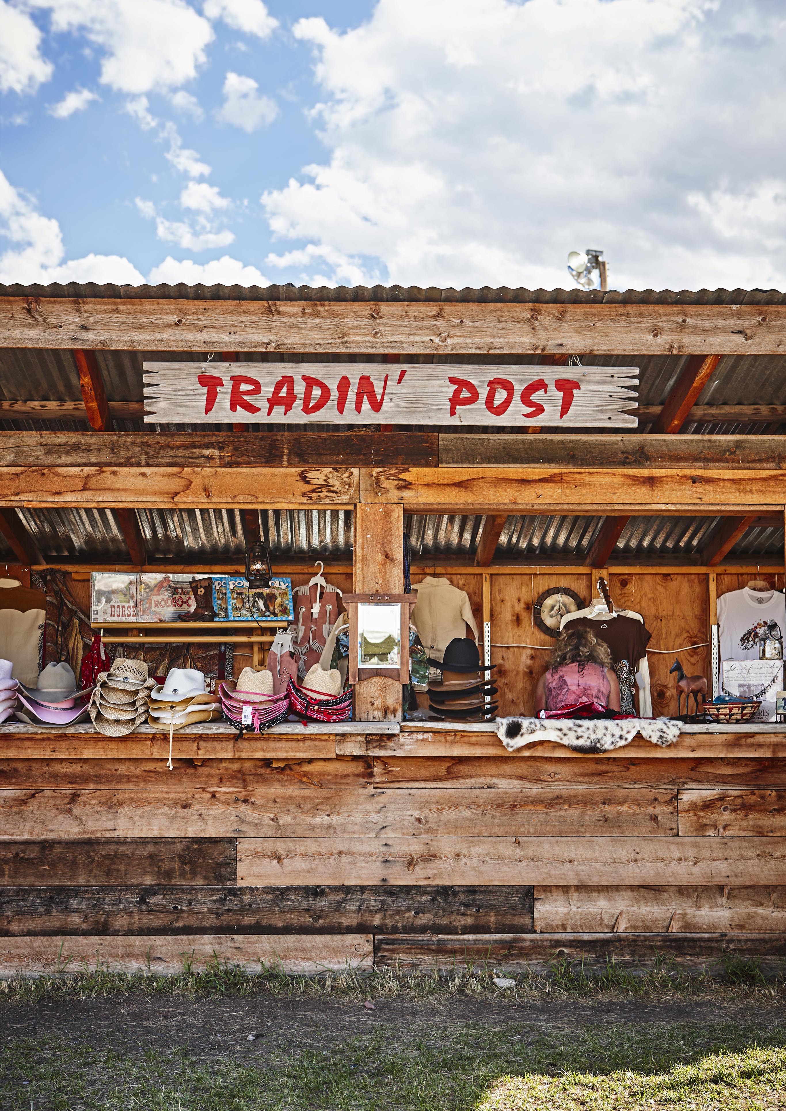 An editorial capture of wooden trading post booth displays cowboy hats, shirts, and various goods under a corrugated metal roof with a sign reading "TRADIN' POST," reminiscent of frontier stops in the Rocky Mountains of North America.