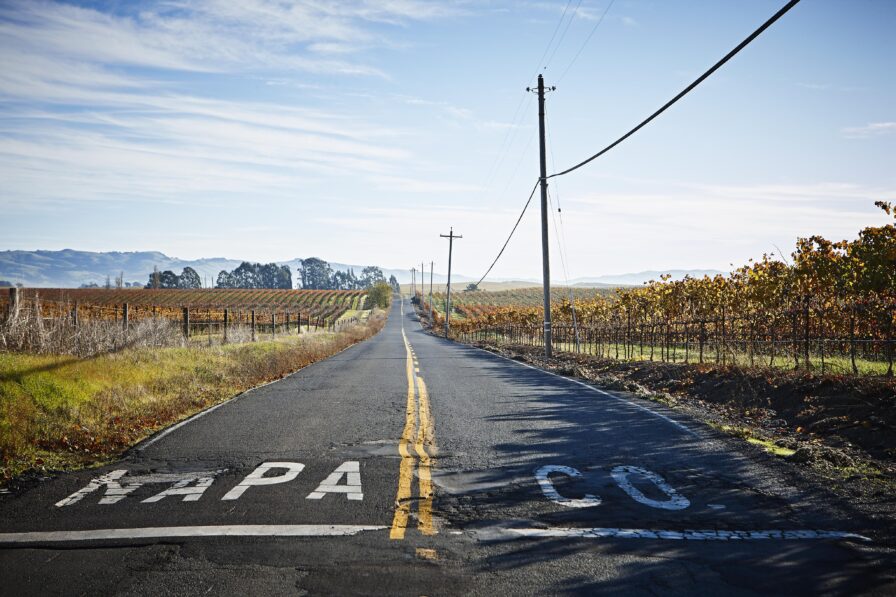 Narrow road with double yellow lines, flanked by vineyards under clear blue sky. Power poles and wires run alongside. "NAPA CO" painted on road surface, indicating Napa County.