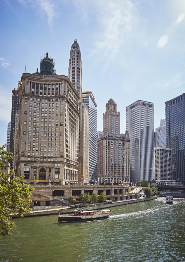 A boat travels on the Chicago River with several tall office buildings and skyscrapers in the background on a sunny day.