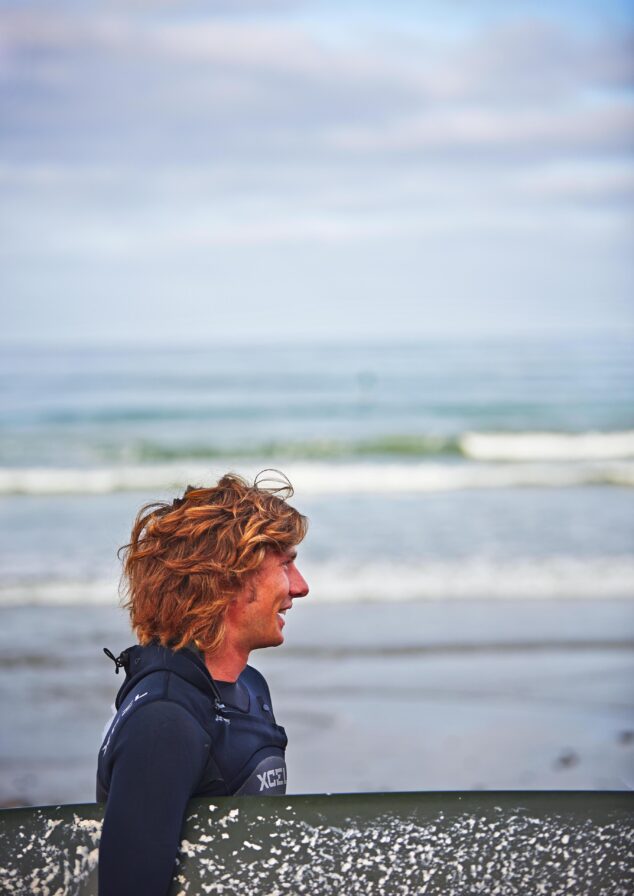 Authentic lifestyle capture of person with wavy hair holding a surfboard, wearing a wetsuit, stands on the beach with waves and cloudy San Diego sky in the background.