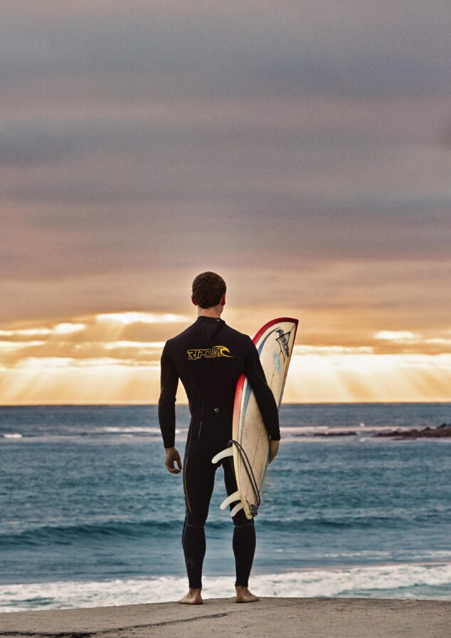 A person in a wetsuit holds a surfboard under one arm, standing on the San Diego shore facing the ocean at sunset under a cloudy sky as rays of sun rush through the cloud cover.