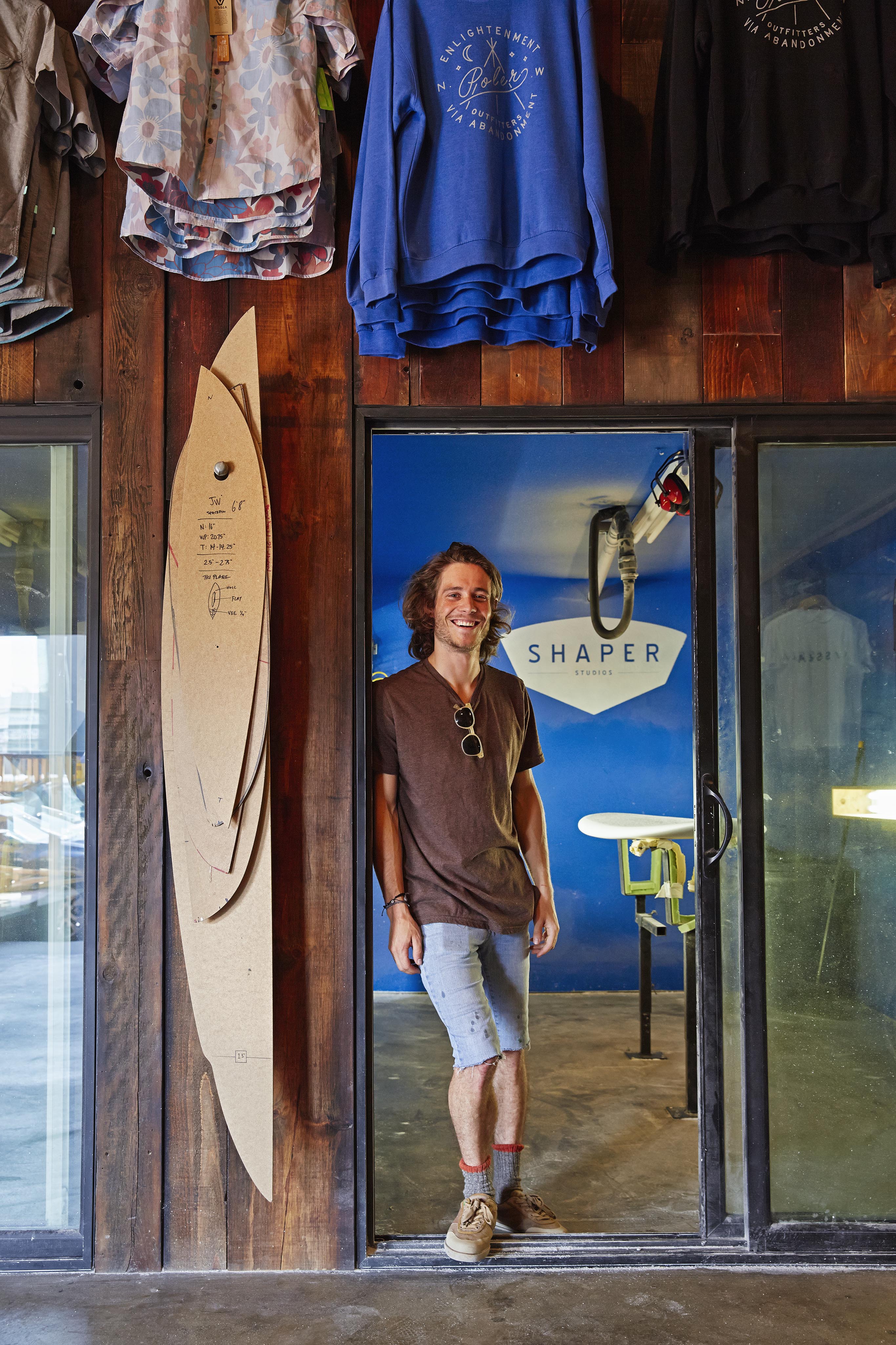 Person smiling in a doorway of a workshop with surfboards and clothing on the wall. A sign reads "Shaper" inside.