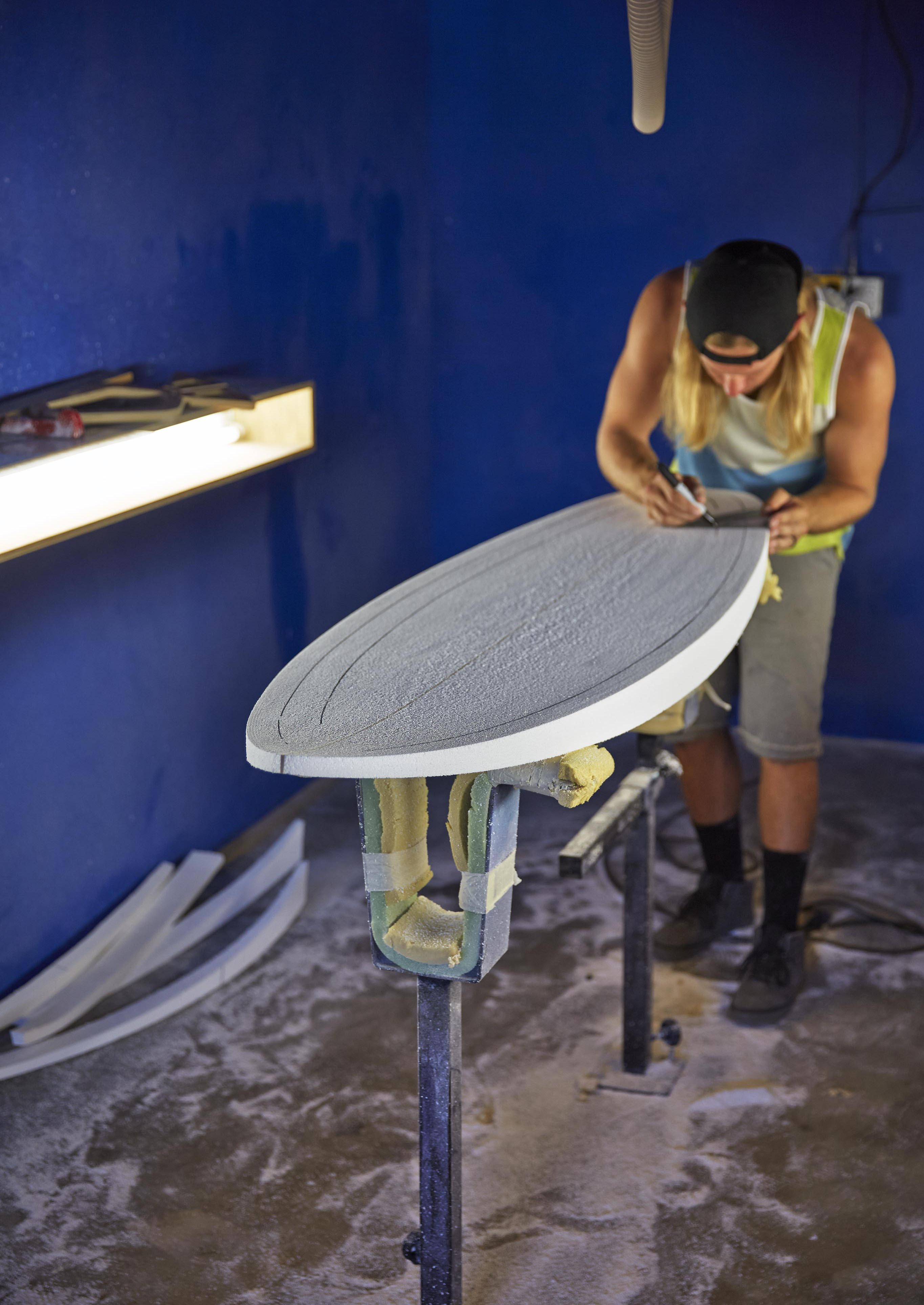 An authentic lifestyle capture of person shaping a surfboard in a San Diego board shop, with deep blue walls and dust covered floors; person leaned over using a tool to refine its contours of the board.
