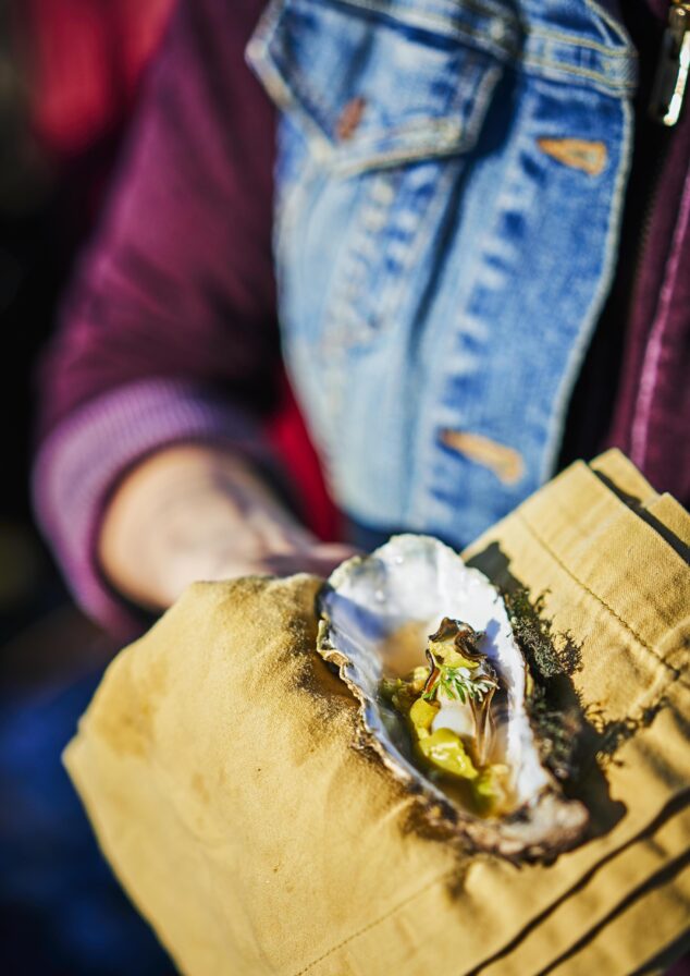 Person holding a close up view of large open oyster on a yellow cloth, with visible garnish and toppings, person wearing a denim and maroon jacket.