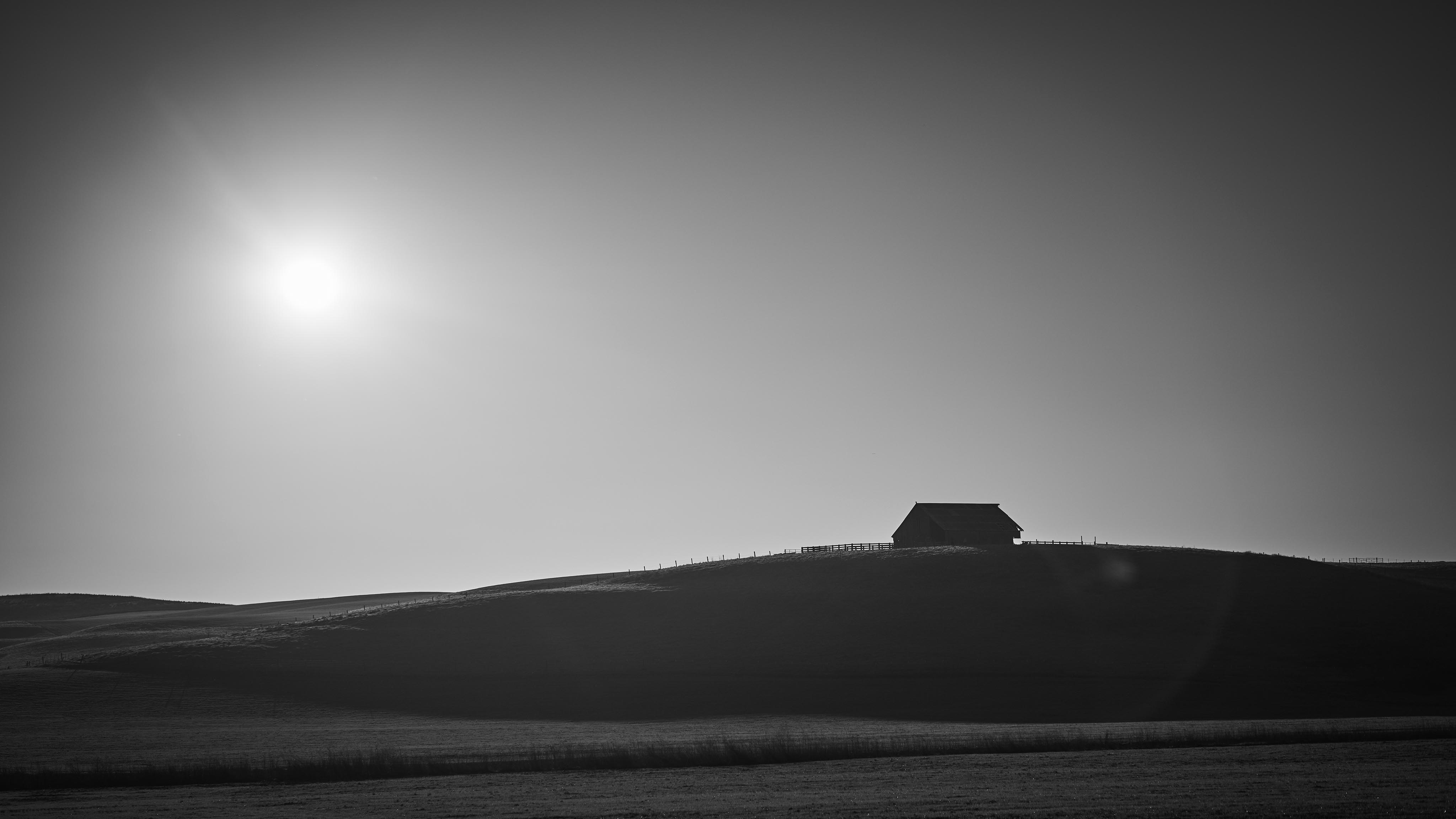 A solitary barn stands on a hill under a bright sun in a black and white landscape.