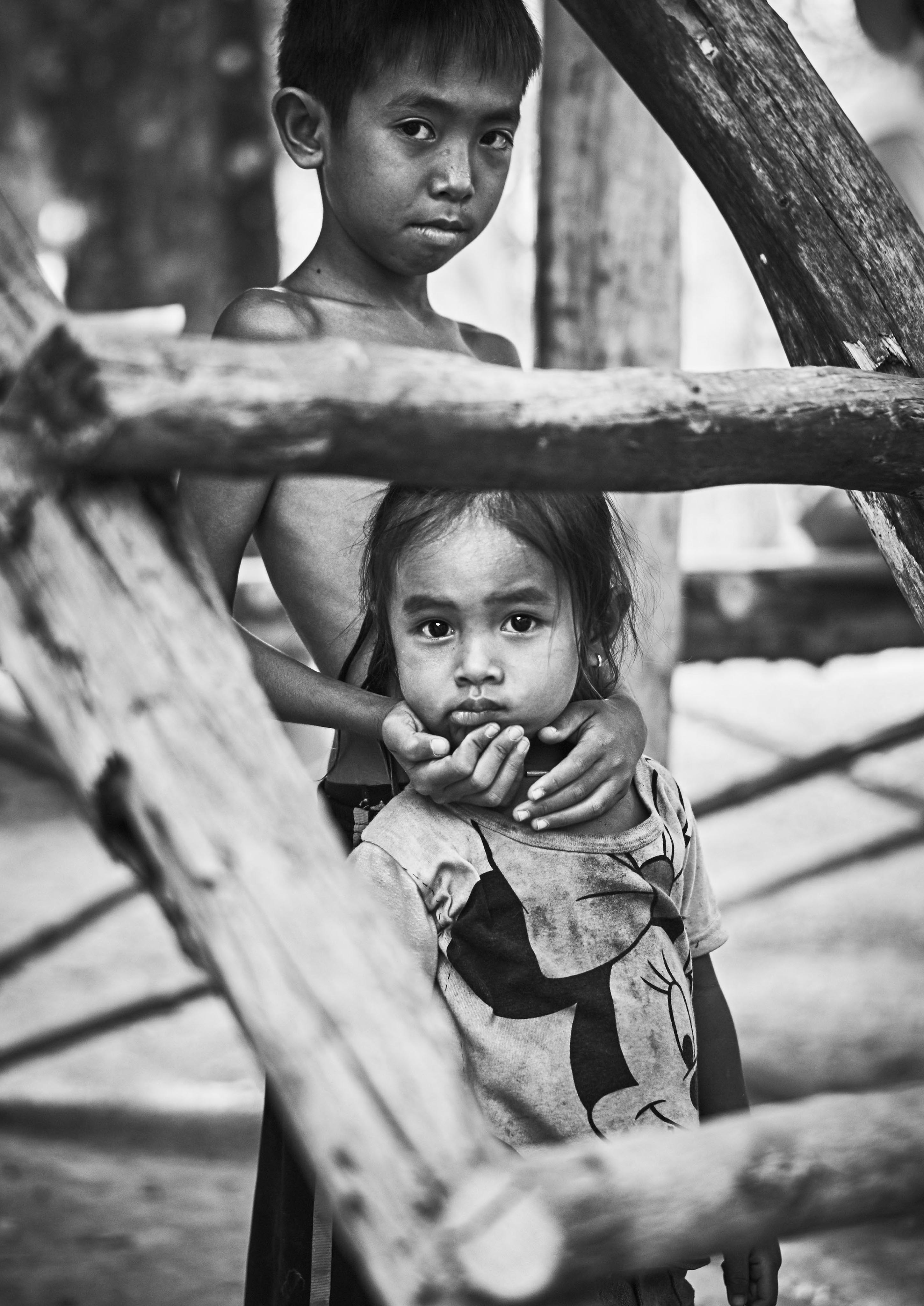 A black and white photo of a young boy holding a younger girl's shoulder, both looking at the camera. Wooden beams frame them in the foreground.