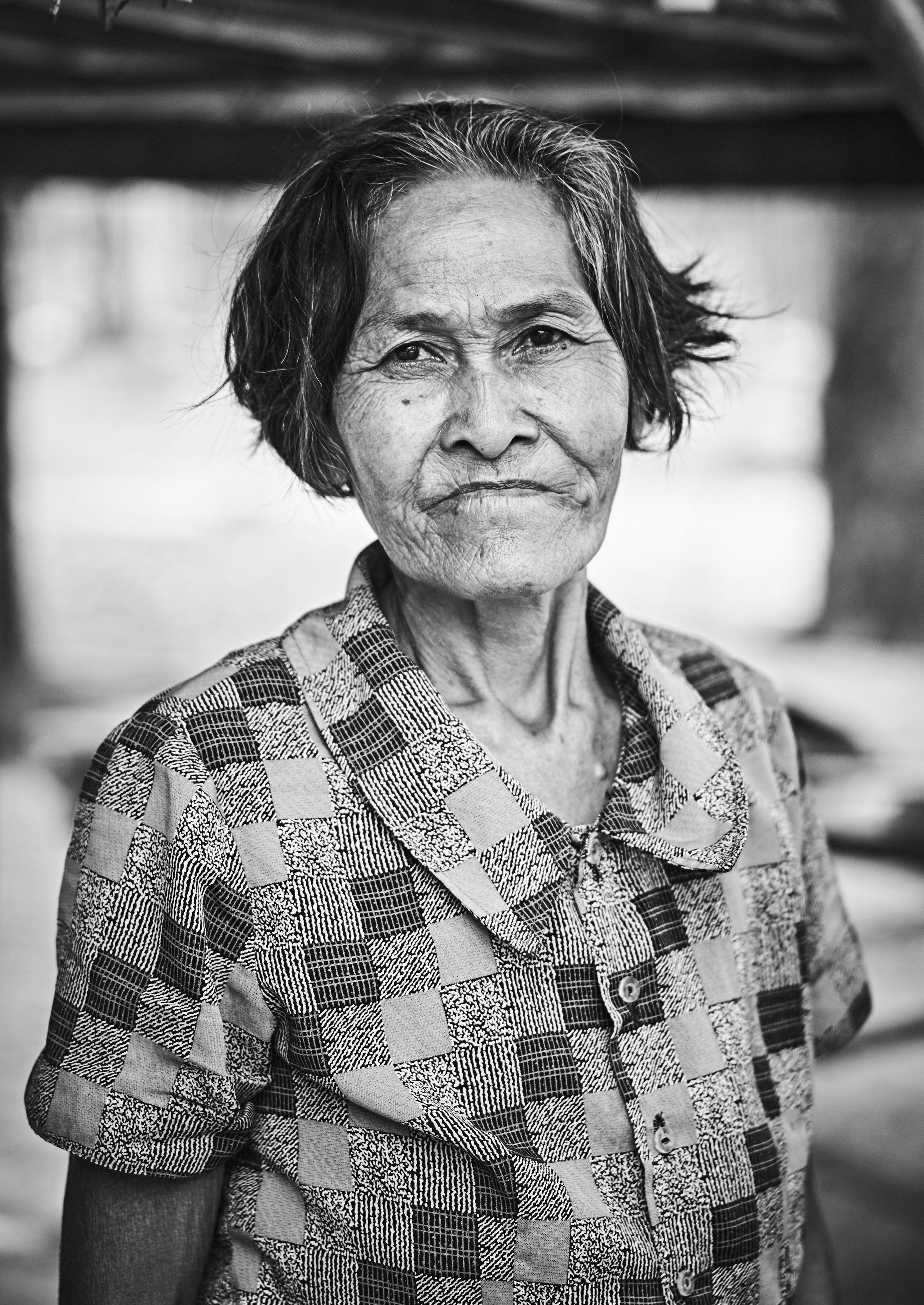 Older woman with short hair, wearing a checkered blouse, standing outdoors with a neutral expression. Black and white photograph.