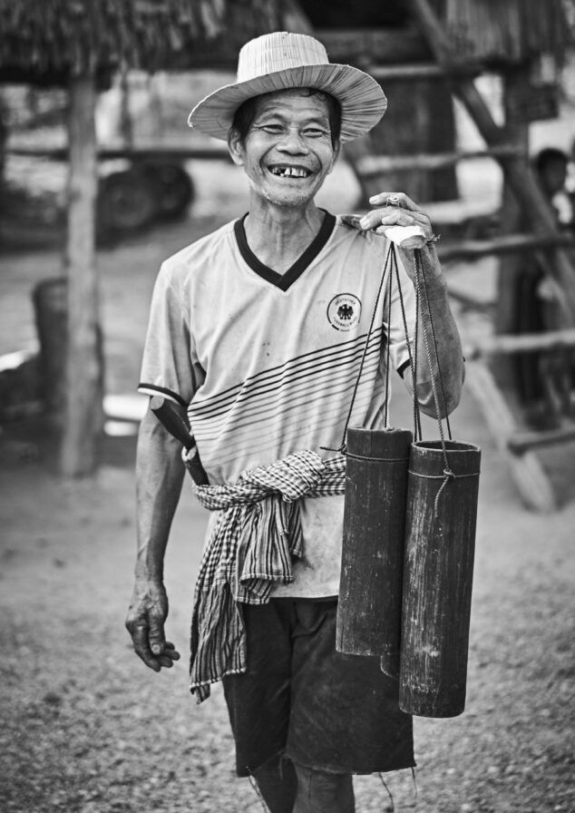 Smiling man in a straw hat carries two cylindrical containers outdoors, wearing a striped shirt and checkered cloth around his waist.