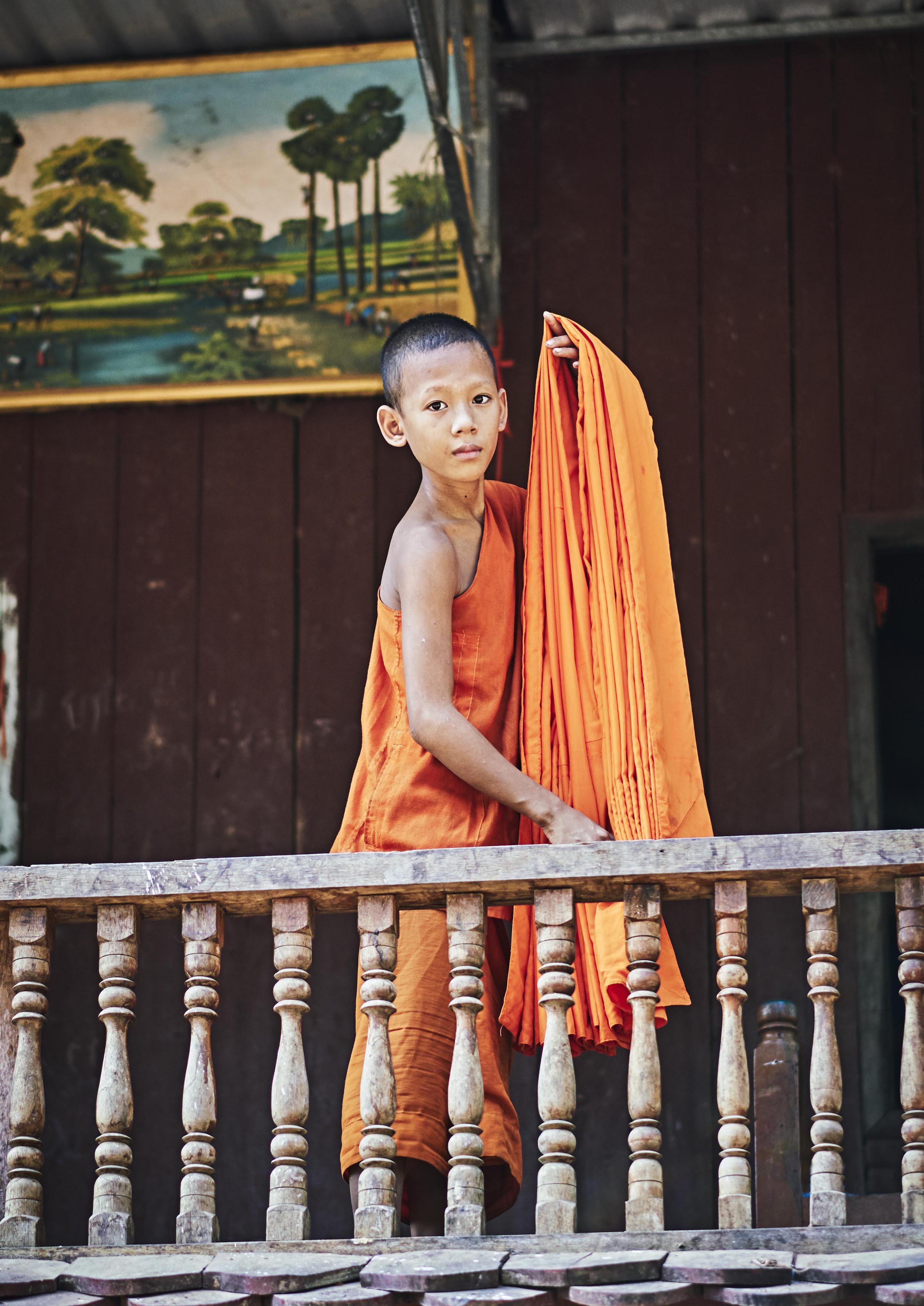 A young monk in an orange robe stands on a wooden balcony, holding additional folded robes. A painting of trees and a lake hangs on the wall behind him.