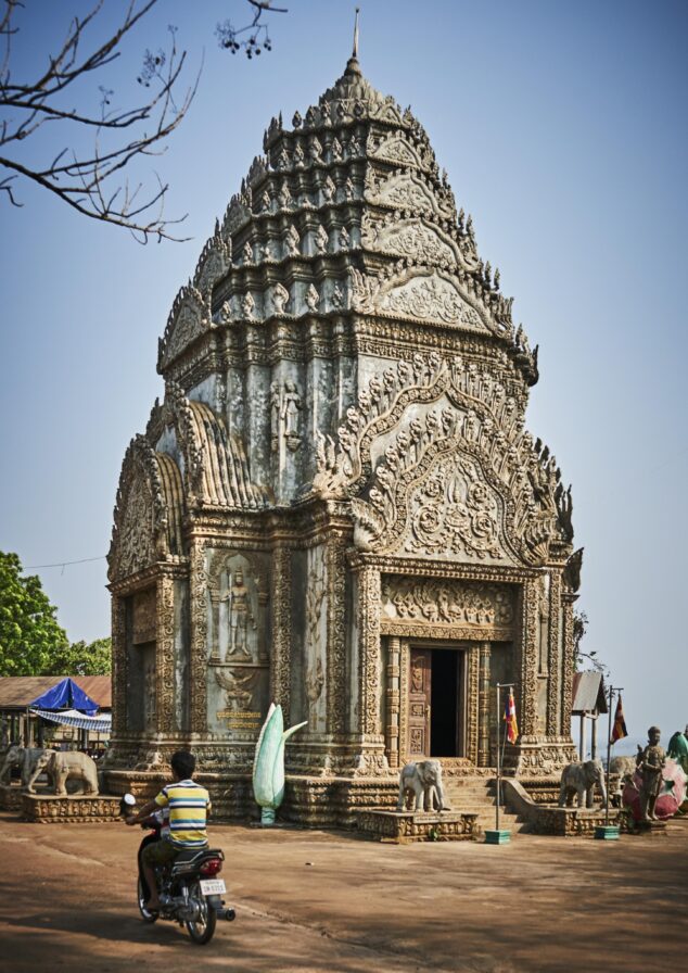 A person on a motorcycle rides past an ornate, intricately carved temple with a pointed roof on a clear day.