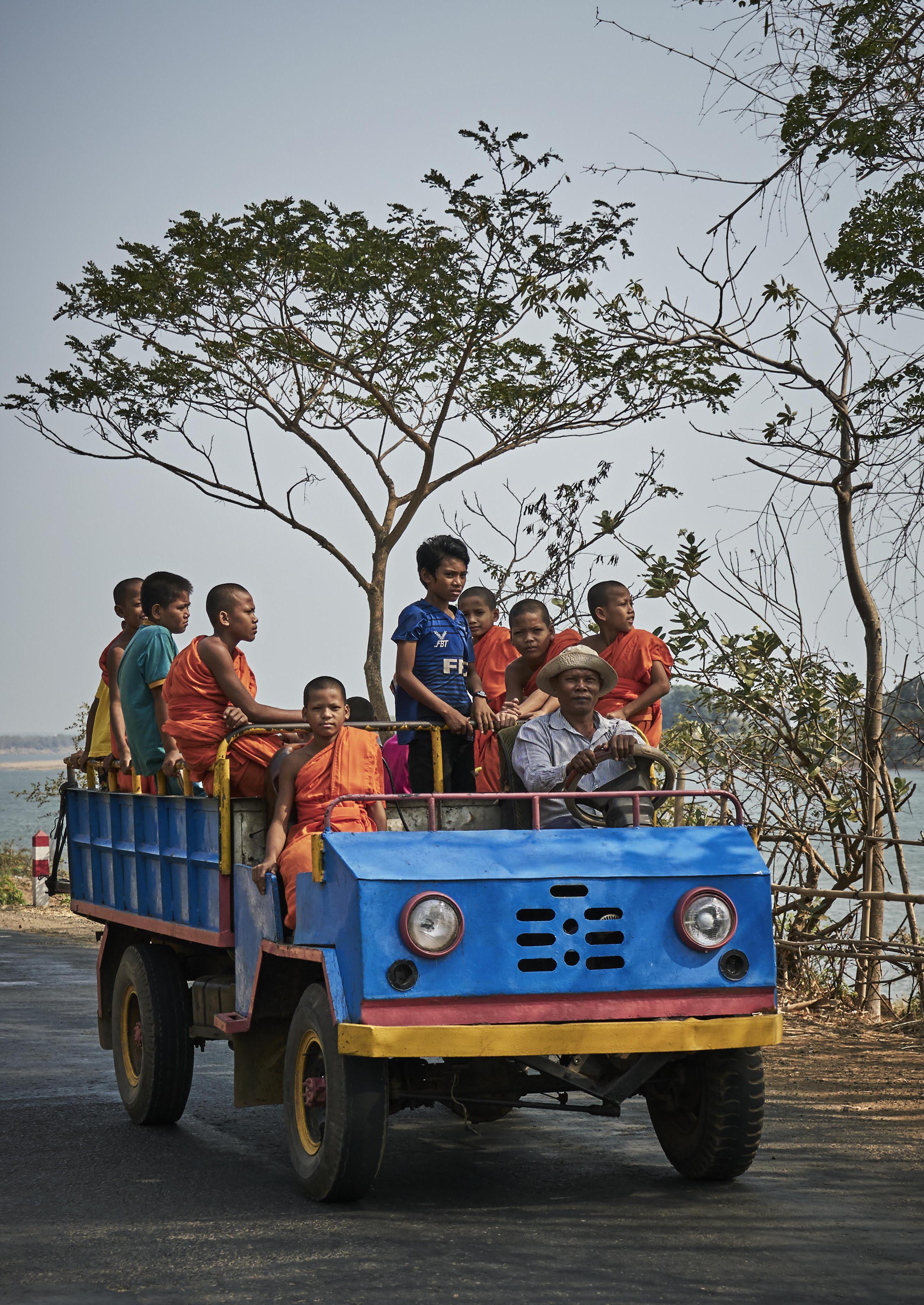 A group of children in colorful clothing ride on the back of a vibrant blue truck driven by a man wearing a hat. Trees are visible in the background on a sunny day.