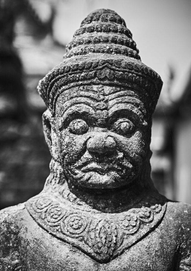 A black and white photo of an ancient, intricately carved stone statue with a detailed face, spiral beard, and a textured headdress, set against a blurred background.