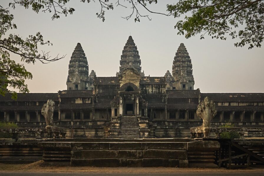 The image shows the front view of Angkor Wat, with its iconic towers and stone facade, surrounded by trees under a clear sky.
