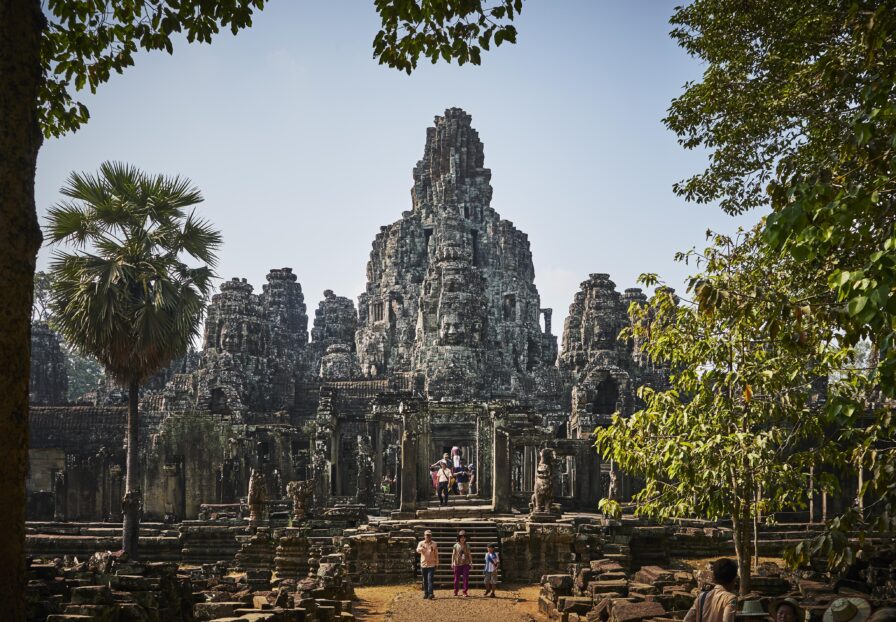 Visitors walk towards the ancient stone temple of Bayon in Angkor, surrounded by trees under a clear sky.
