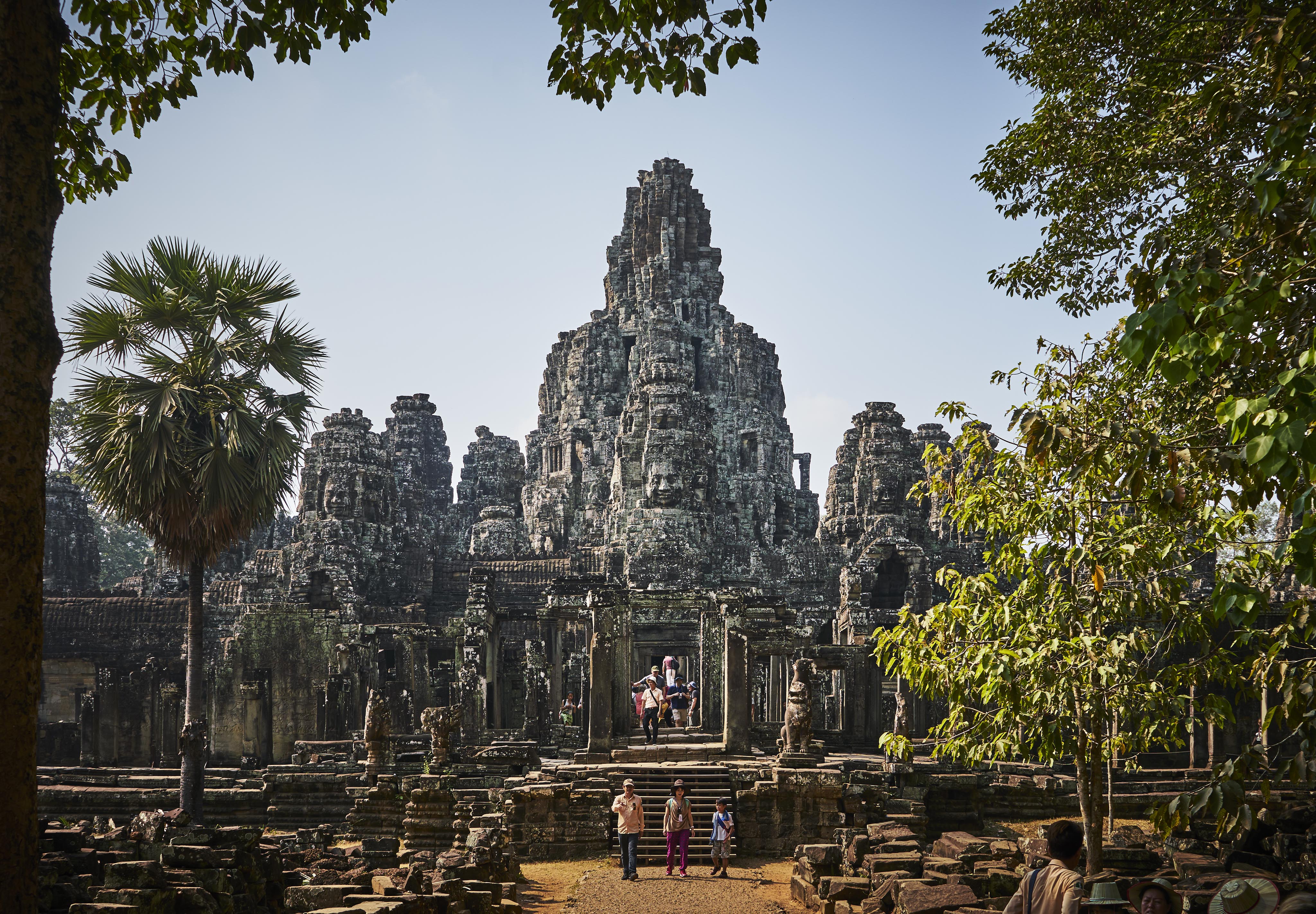 Visitors walk towards the ancient stone temple of Bayon in Angkor, surrounded by trees under a clear sky.