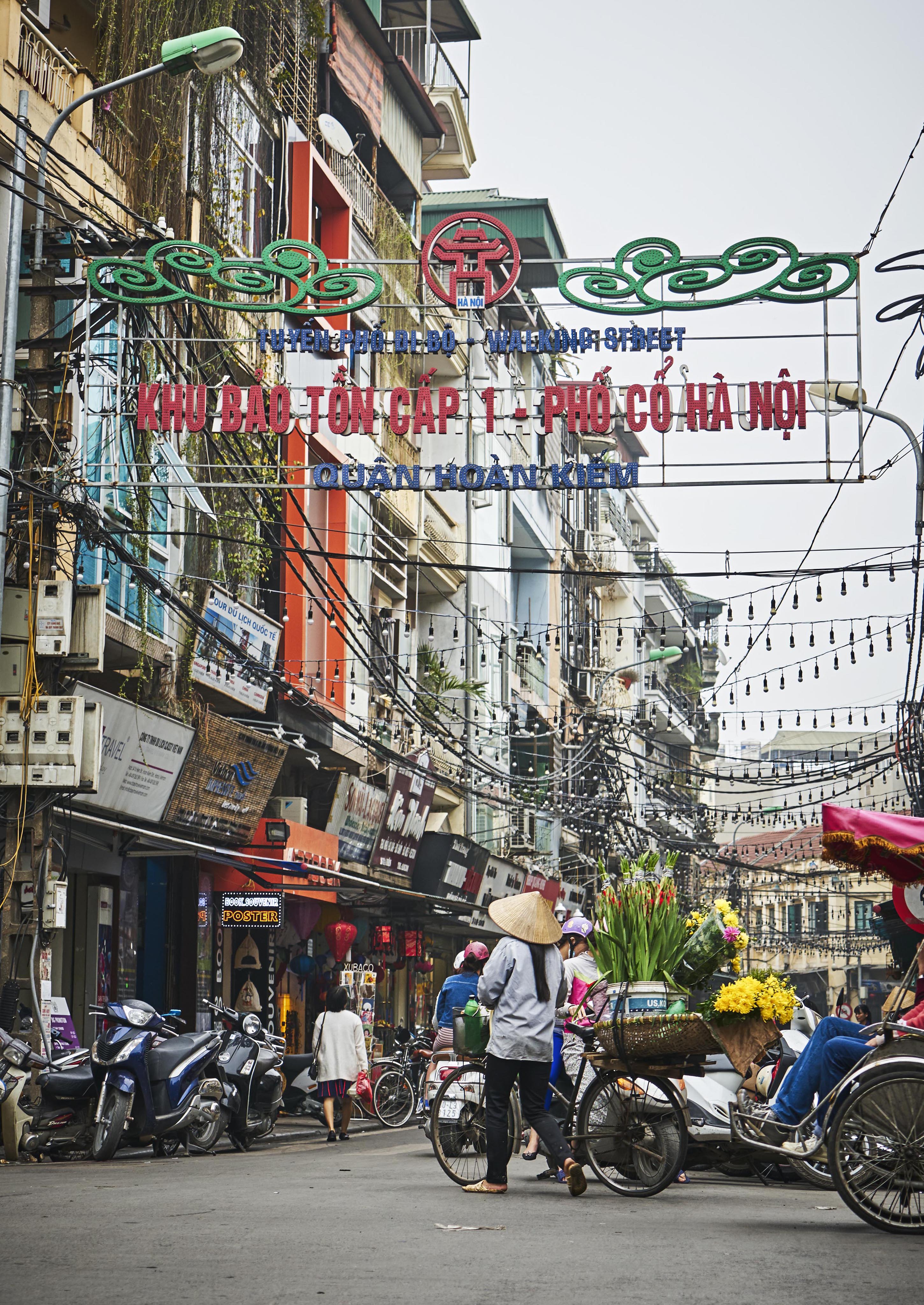 A busy street in Hanoi's Old Quarter, with people walking and riding motorcycles. A person on a bicycle carries flowers. A large sign in Vietnamese hangs above the road.