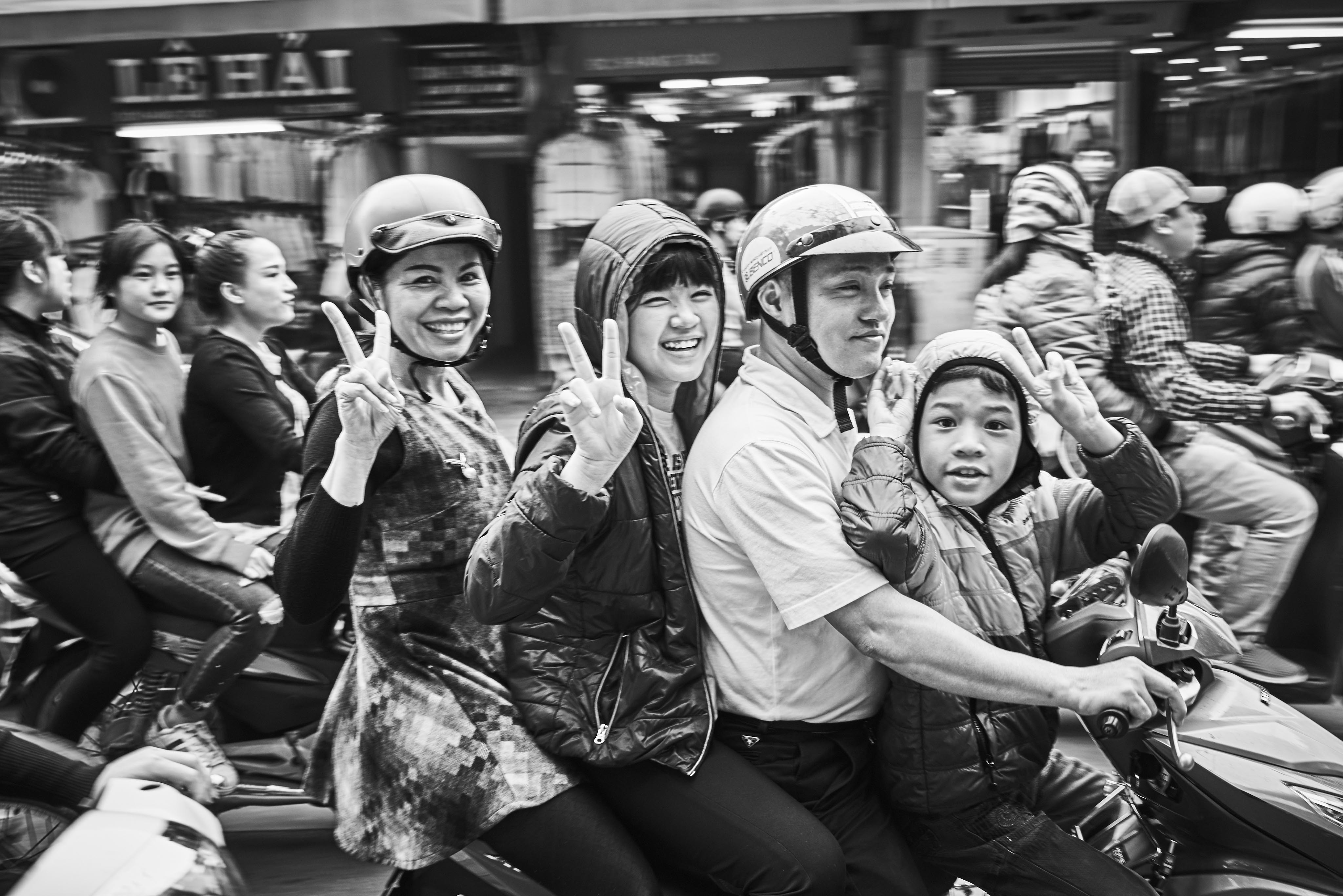 A group of people, two women and a man with a child, ride on a crowded street. They wear helmets and make peace signs while sitting on a motorcycle. The background is bustling with traffic.
