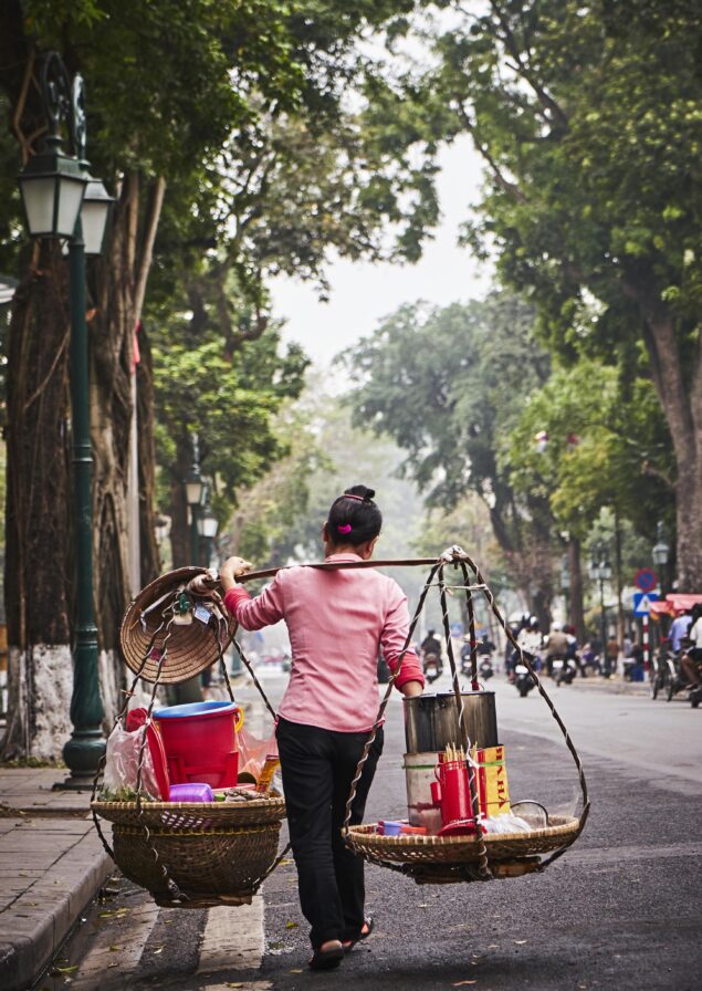 A person carries a traditional bamboo pole with baskets filled with various items, walking along a tree-lined street.