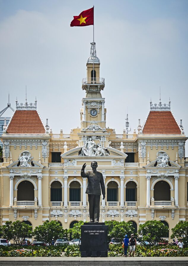 Statue of a man in front of a yellow colonial-style building with a clock tower. A red flag with a yellow star flies above. People are visible near the statue.