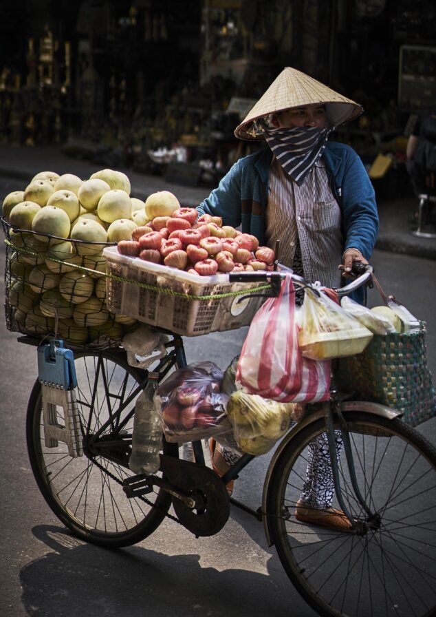 A street vendor wearing a conical hat pushes a bicycle loaded with baskets of apples, melons, and other goods.