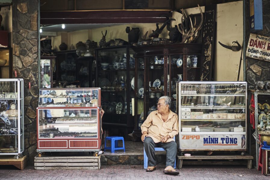 A man sits on a blue chair outside a shop displaying various items in glass cases.