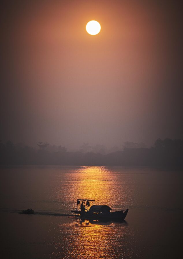 A boat travels on a calm river against the backdrop of a setting sun, casting a warm reflection on the water.