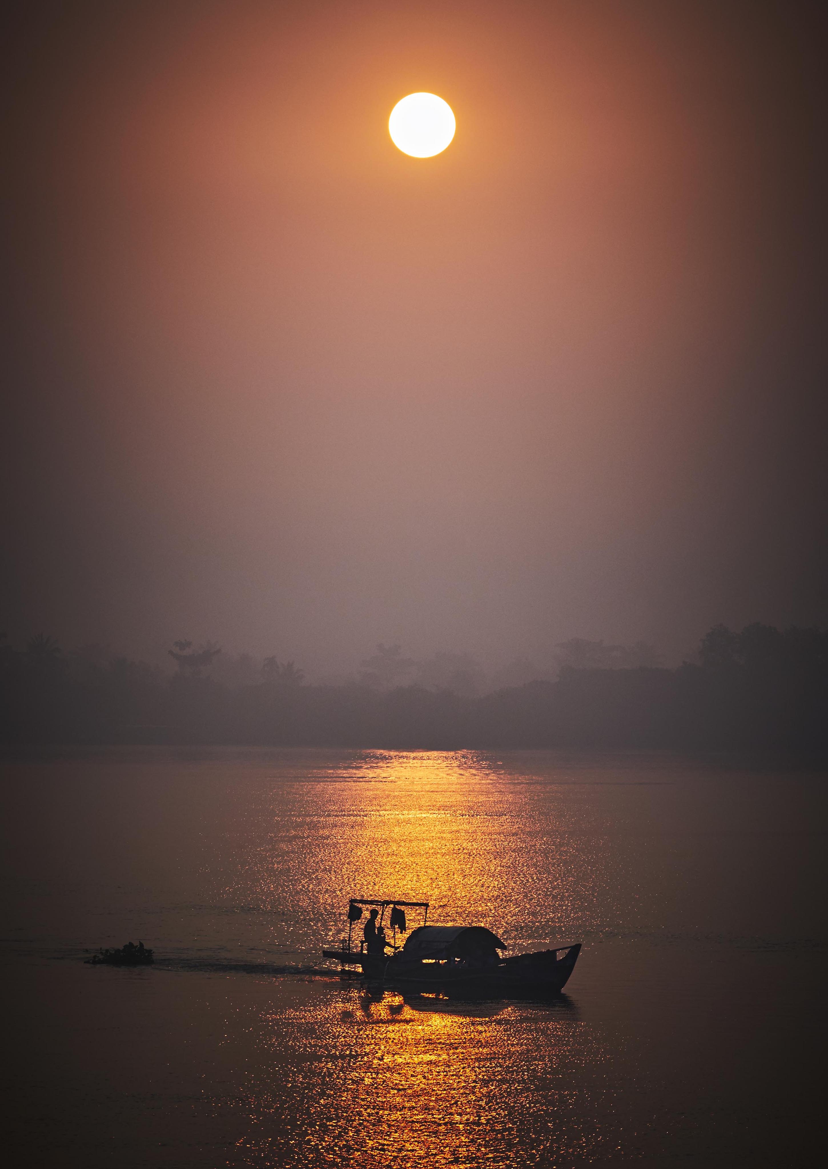A boat travels on a calm river against the backdrop of a setting sun, casting a warm reflection on the water.
