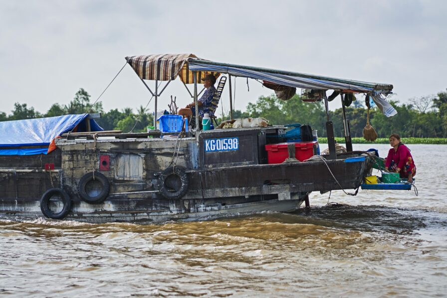 A weathered boat with a canopy travels on a river, carrying a person at the front and another seated near the back. The vessel displays identification KG0158B, surrounded by water and trees.