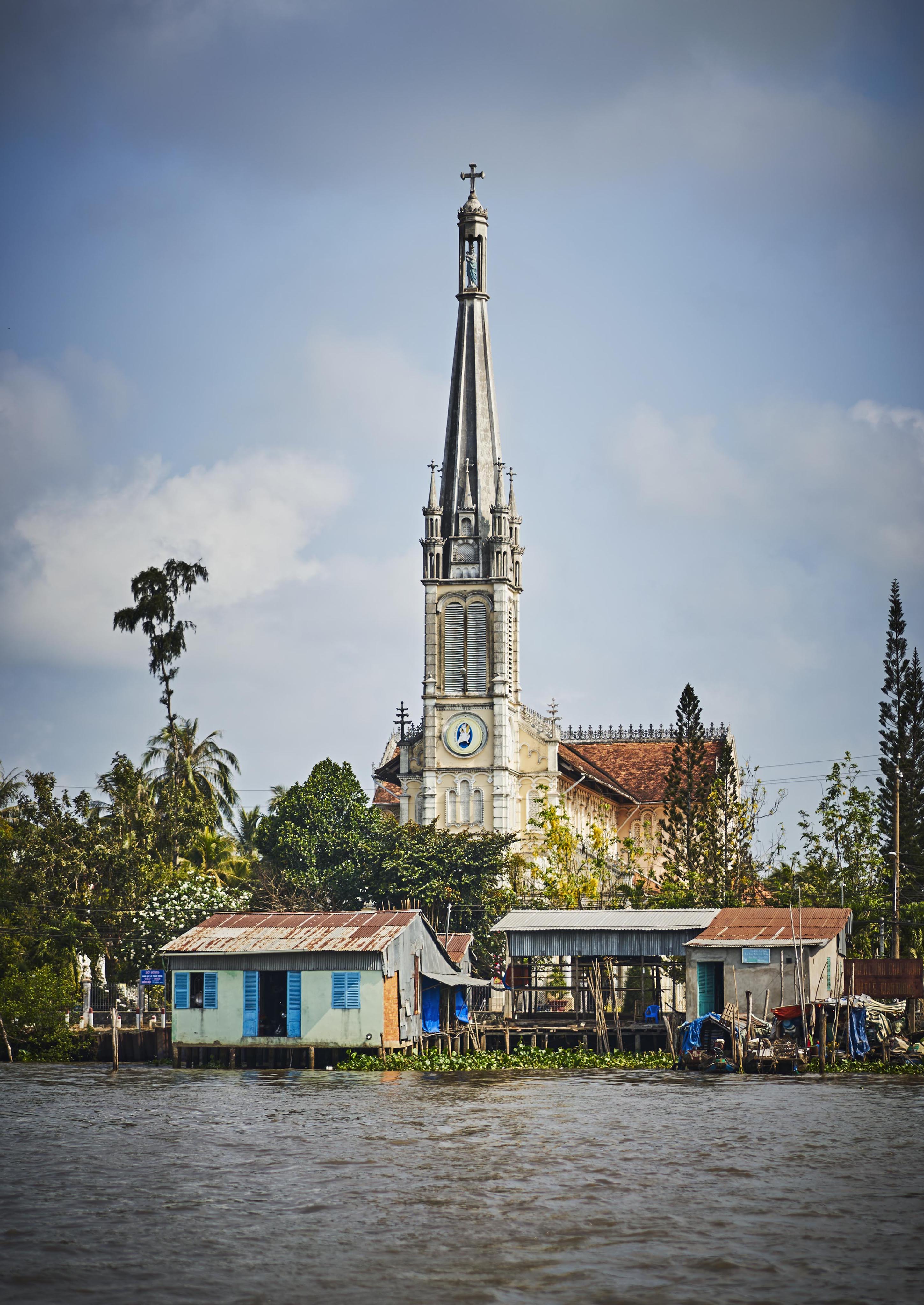 A tall church with a clock tower stands behind small houses by a riverbank, under a cloudy sky.