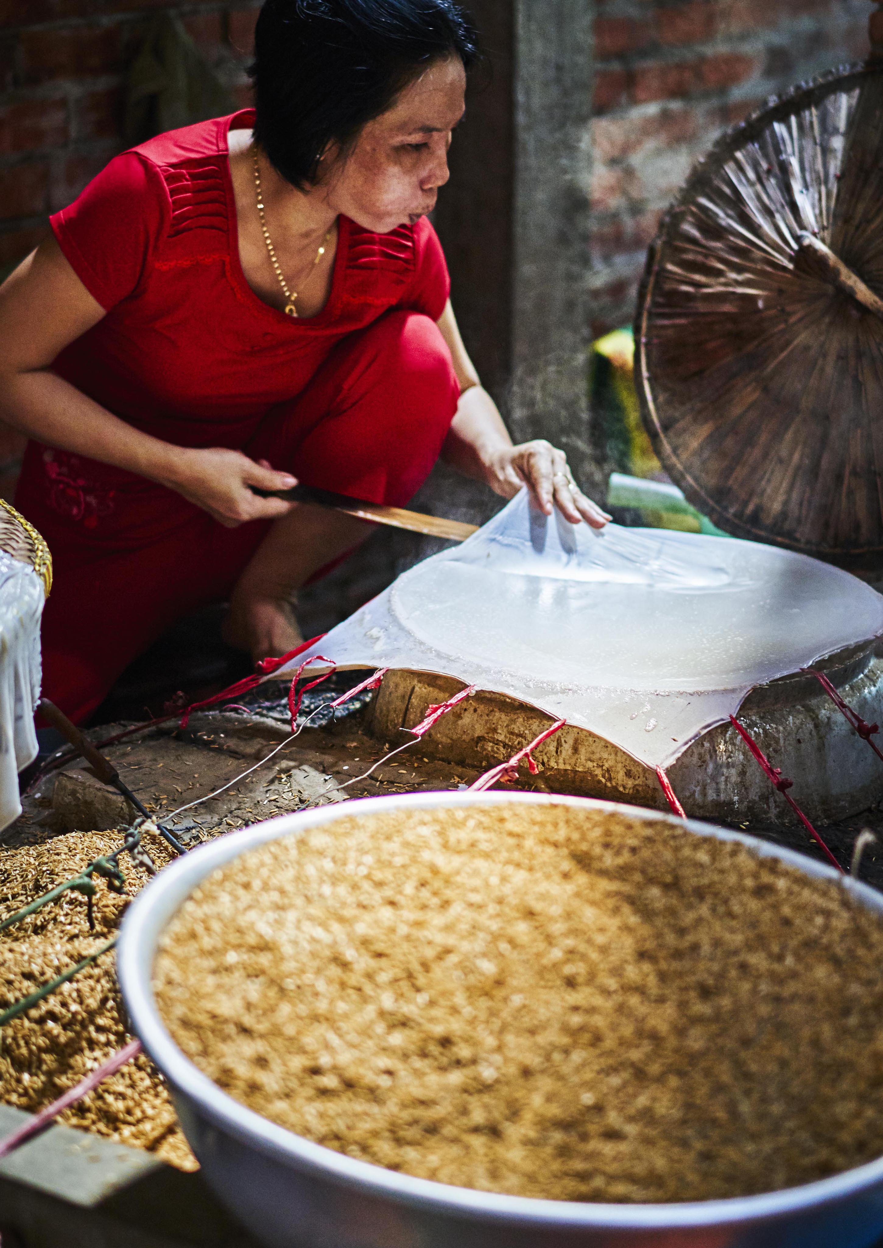 A person in a red outfit making traditional rice paper on a circular frame, with a bowl of grains nearby.