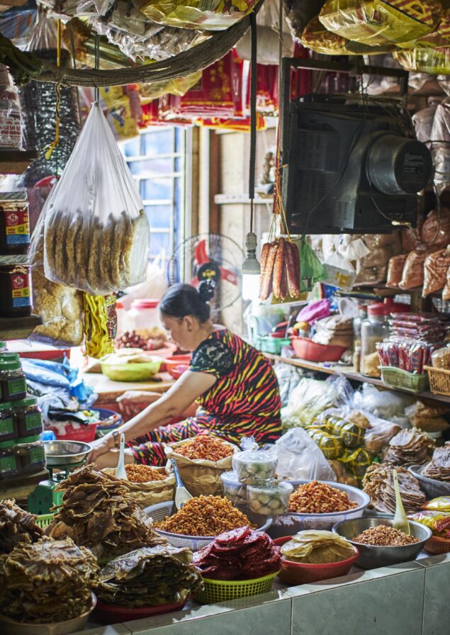 A woman sits surrounded by baskets of various dried foods and spices in a busy market stall filled with colorful packages and jars.