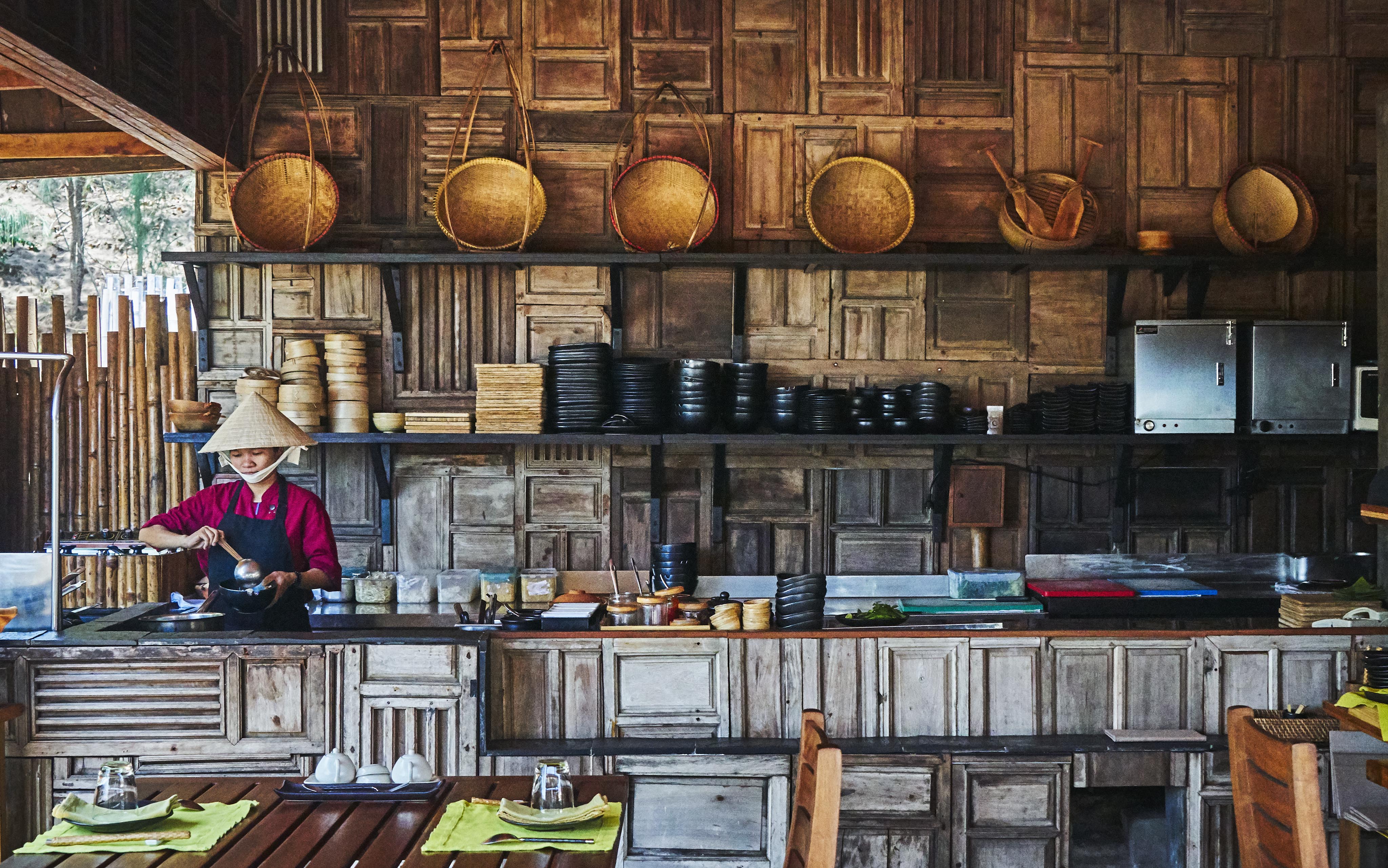 A person wearing a conical hat cooks in a rustic kitchen with wooden decor and shelves filled with baskets and dishes.