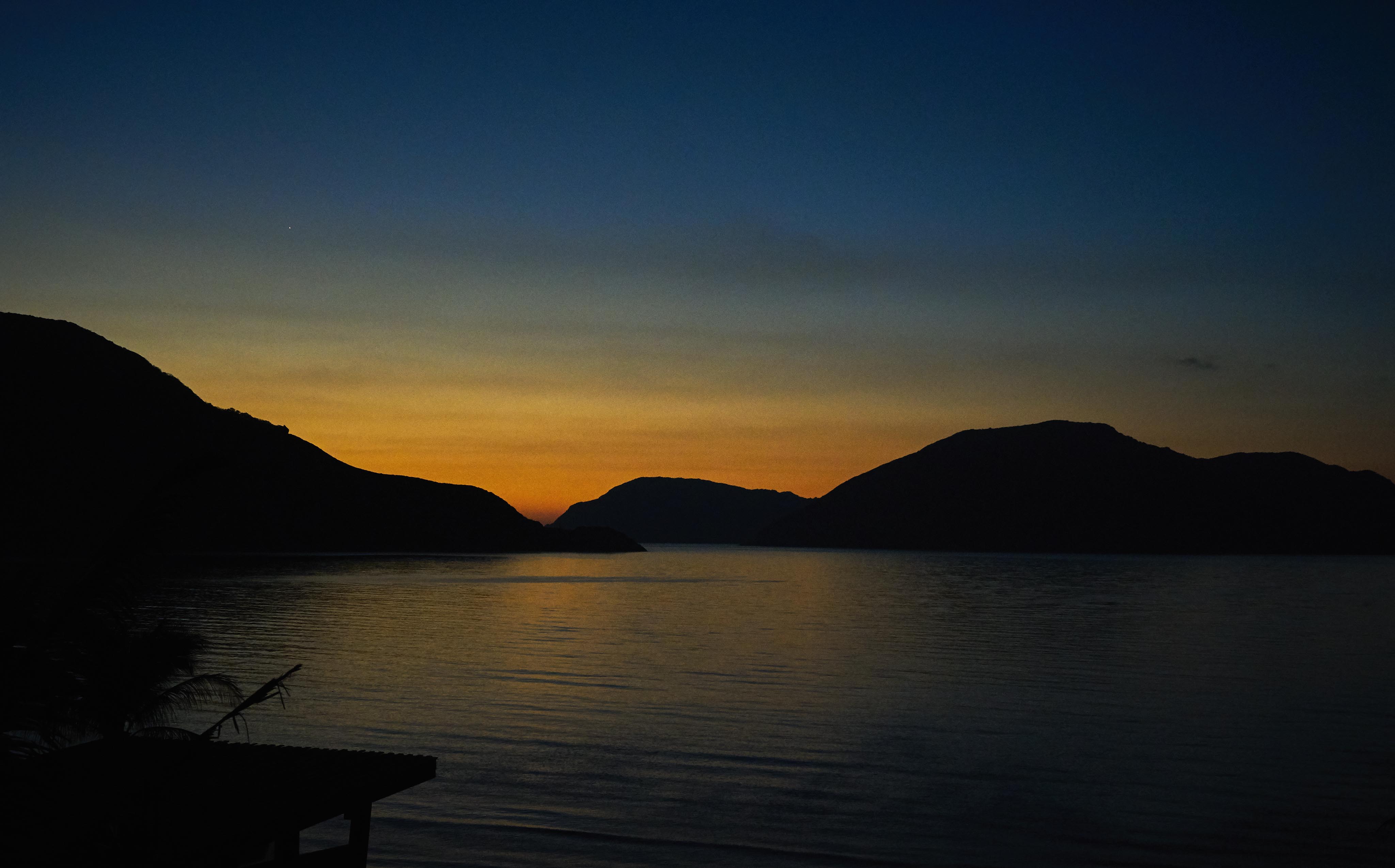 Sunset over a calm body of water with silhouetted hills in the background and a dark foreground.