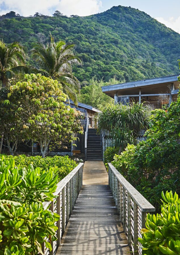 A wooden walkway leads through lush greenery toward a building, with a densely forested mountain in the background.