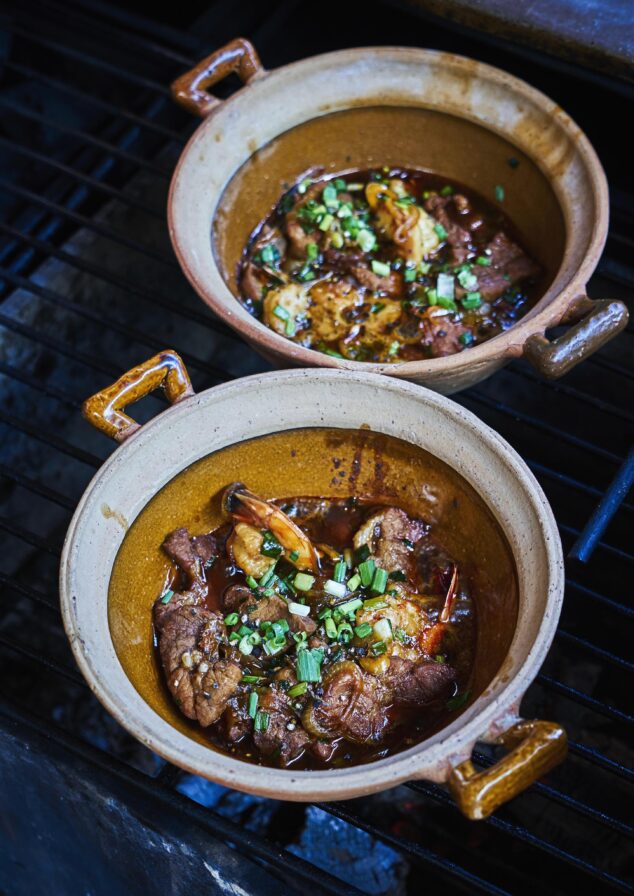 Two clay pots on a grill contain meat dishes with sauce, garnished with chopped green onions.