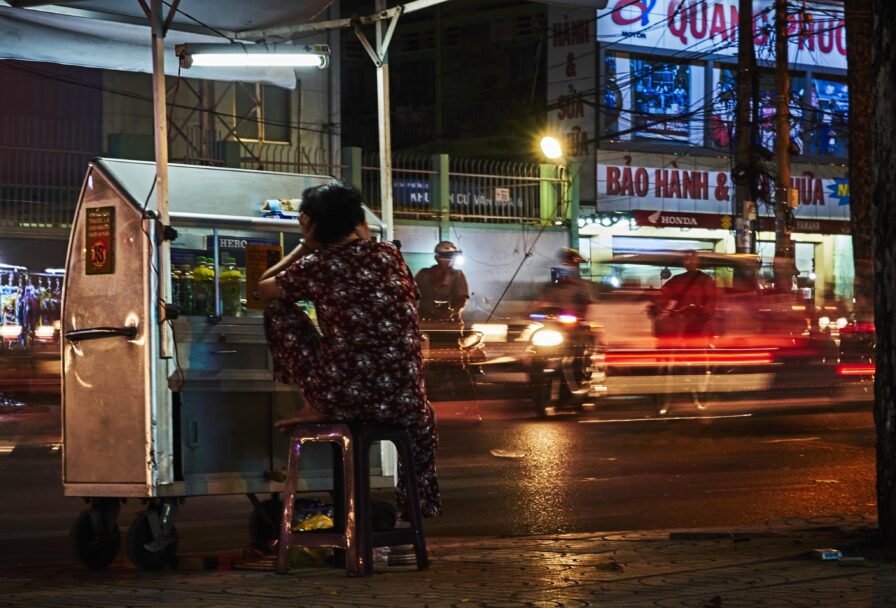 A person in patterned clothing sits on a stool at a nighttime street stall, facing away, while blurred traffic moves quickly past on a busy urban road.