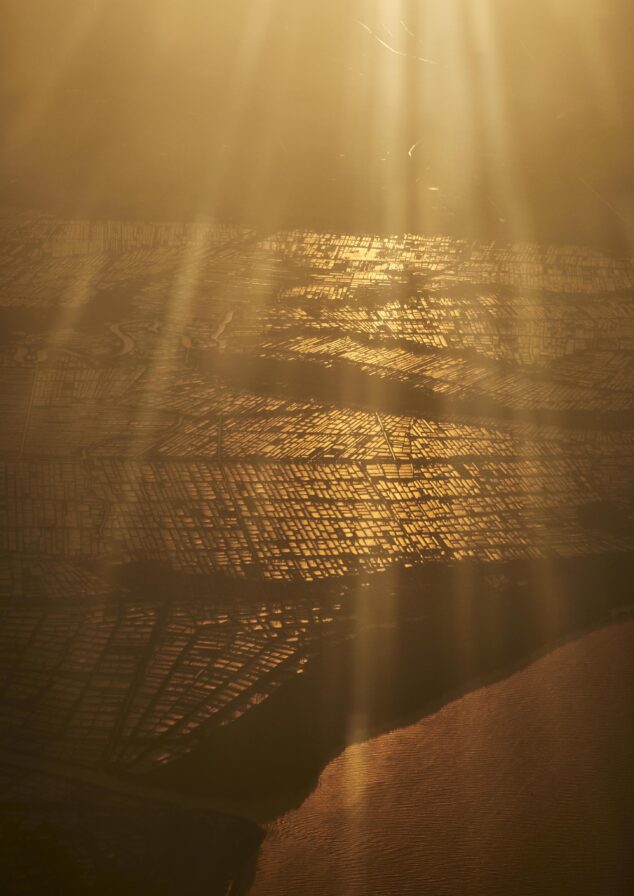 Aerial view of a city grid bathed in golden light, with sunlight streaming down. A body of water is visible at the bottom of the image.