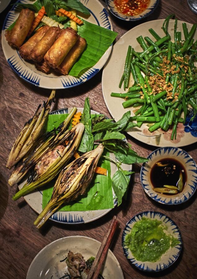 Plates of food on a wooden table include spring rolls, grilled vegetables, green beans, and various sauces.