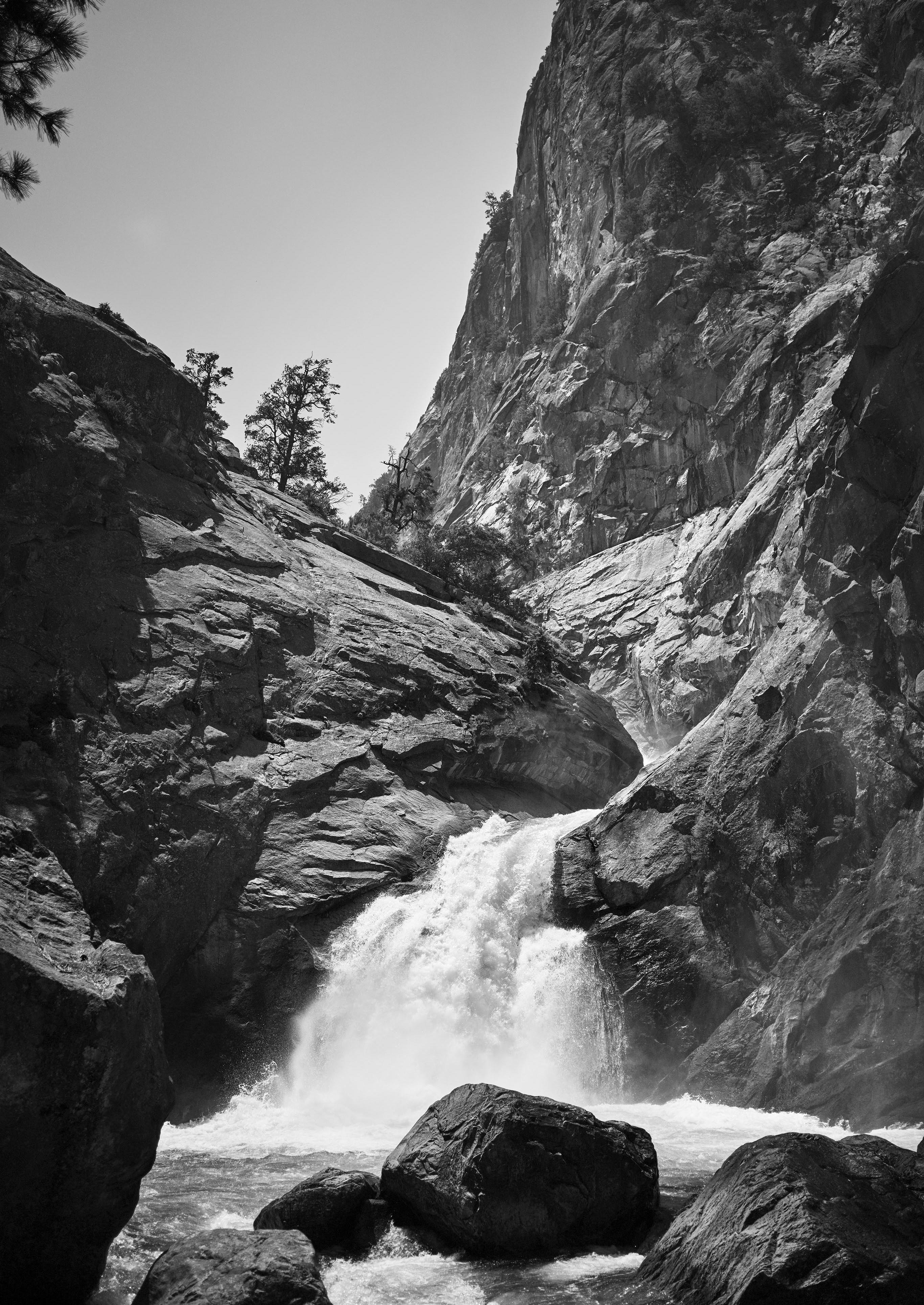 A waterfall cascades down rocky Kings Canyon cliffs surrounded by vast mountains and sparse trees under a clear sky.