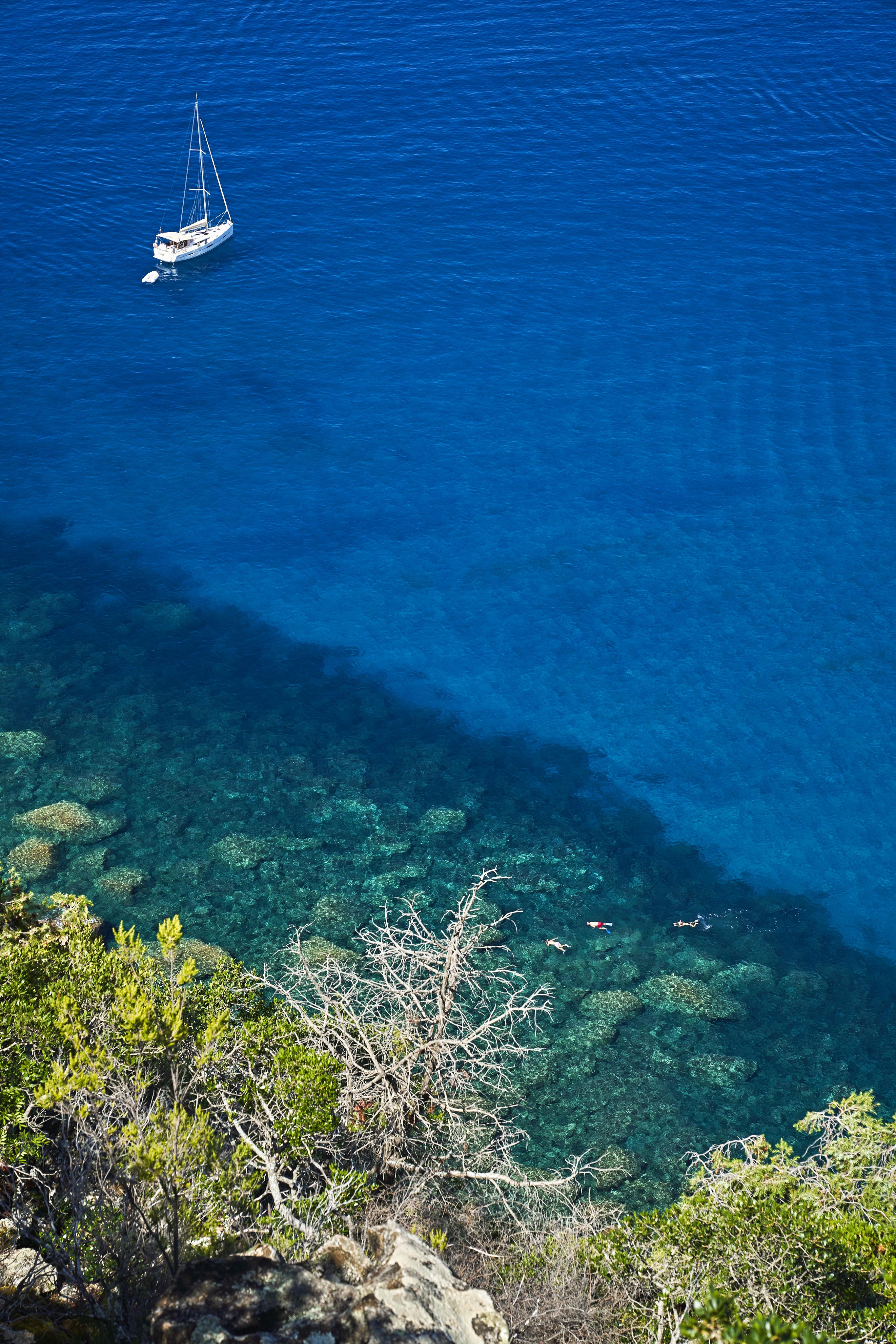 Aerial view of a sailboat anchored in clear blue water near a rocky shoreline with visible vegetation and coral reefs.