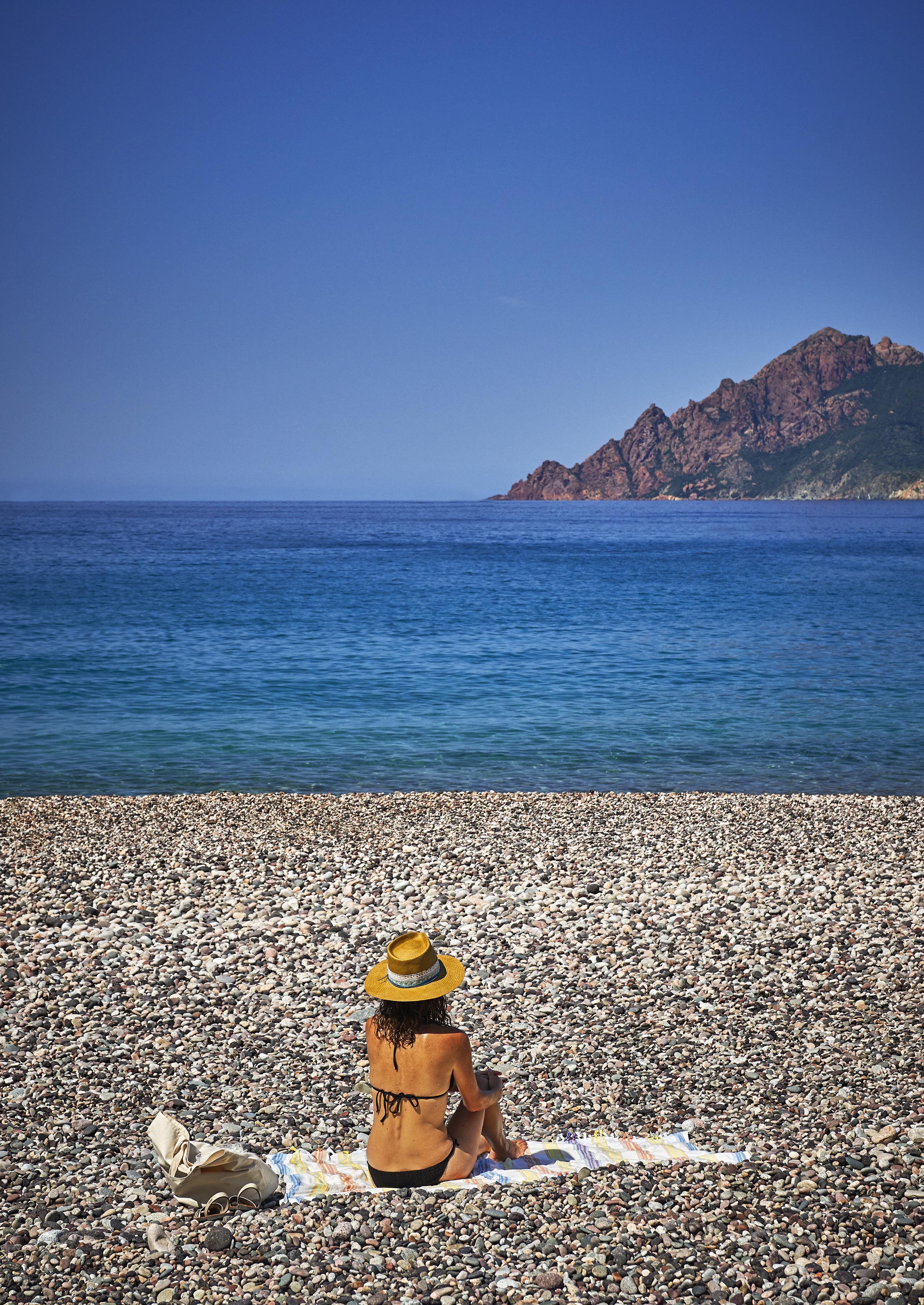 Person in a straw hat and swimwear sits on a pebble beach, facing the calm sea. Mountains are visible in the background under a clear blue sky.