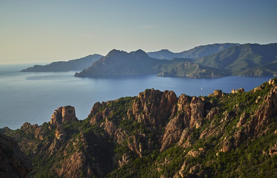 A landscape of rugged mountains and lush greenery overlooking a calm body of water under a clear sky.