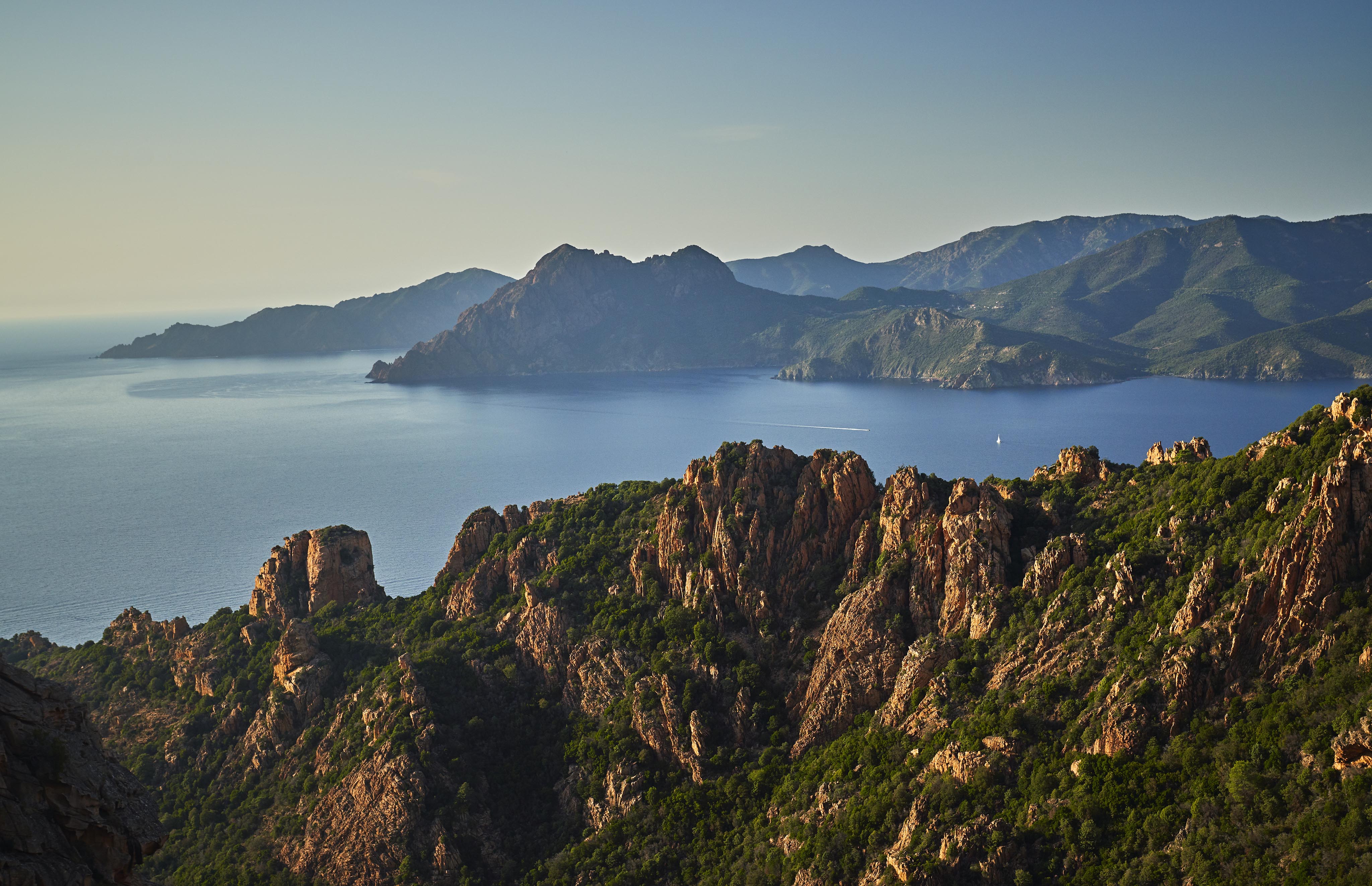 A landscape of rugged mountains and lush greenery overlooking a calm body of water under a clear sky.