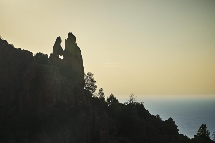 Silhouetted rock formation with a heart-shaped hole overlooks the sea at sunset, surrounded by trees and shrubbery.