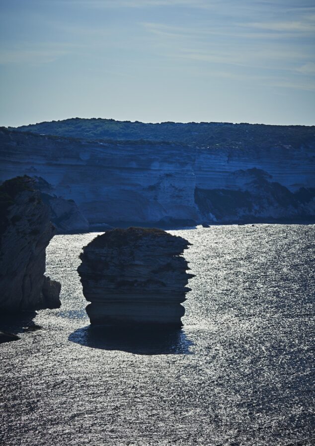 Rock formation surrounded by water under a clear sky, with distant cliffs in the background.