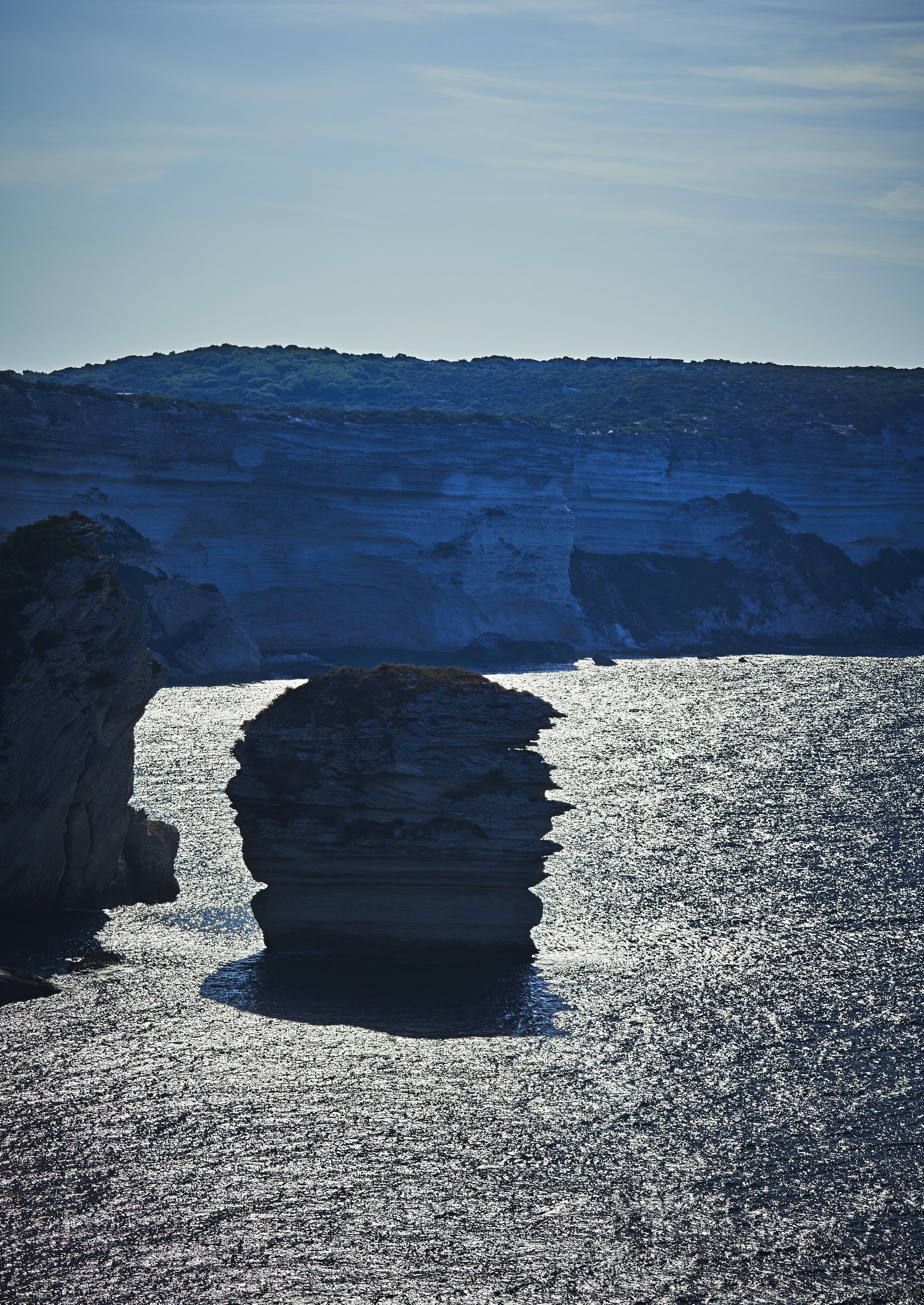 Rock formation surrounded by water under a clear sky, with distant cliffs in the background.