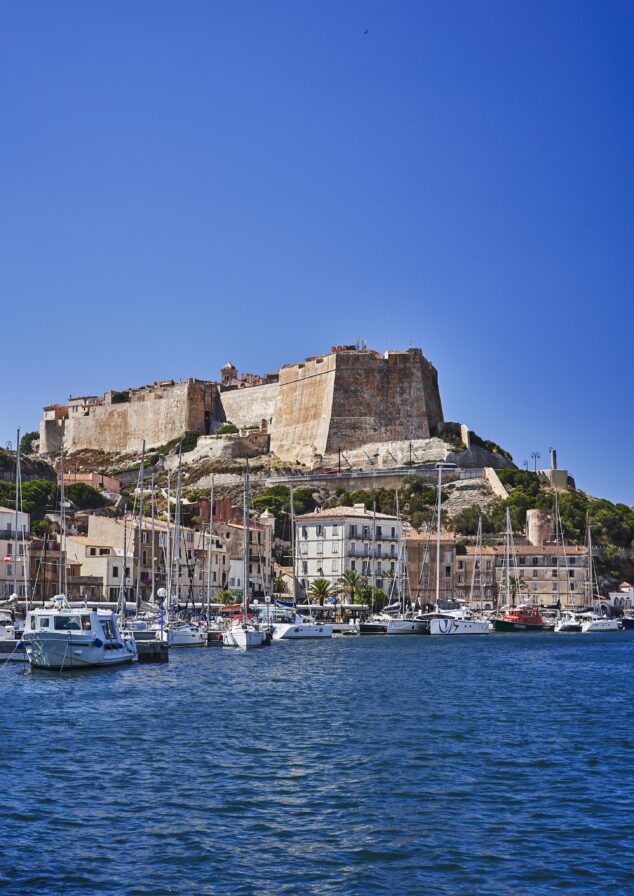 Fortified castle atop a hill overlooking a harbor filled with boats, with a town nestled below under a clear blue sky.