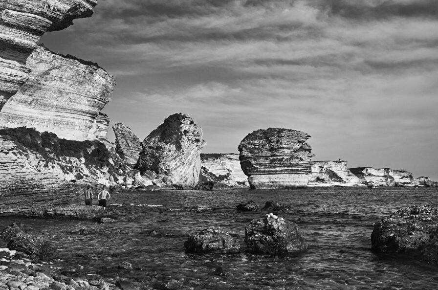 Rocky coastal landscape with large, eroded cliffs and scattered rocks in the shallow water. Two people stand near the shore under a partly cloudy sky. Black and white photo.